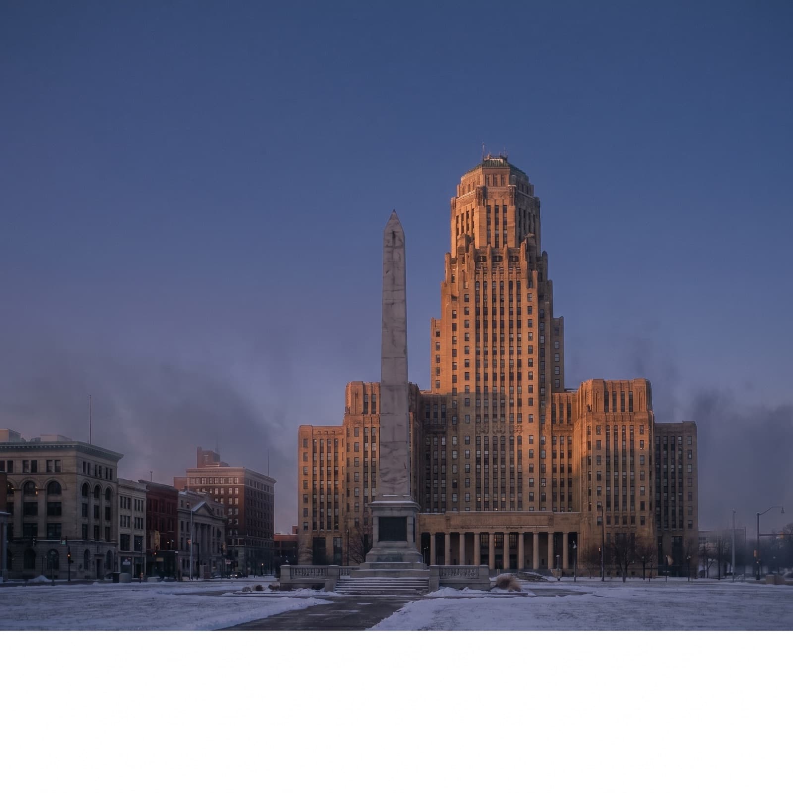 Buffalo, NY — Buffalo City Hall's Art Deco tower rising over Niagara Square at dusk, limestone facade catching the last warm light, McKinley Monument obelisk in the foreground, a dusting of late snow on the steps, no people, no cars, the winter sky going deep blue behind it.