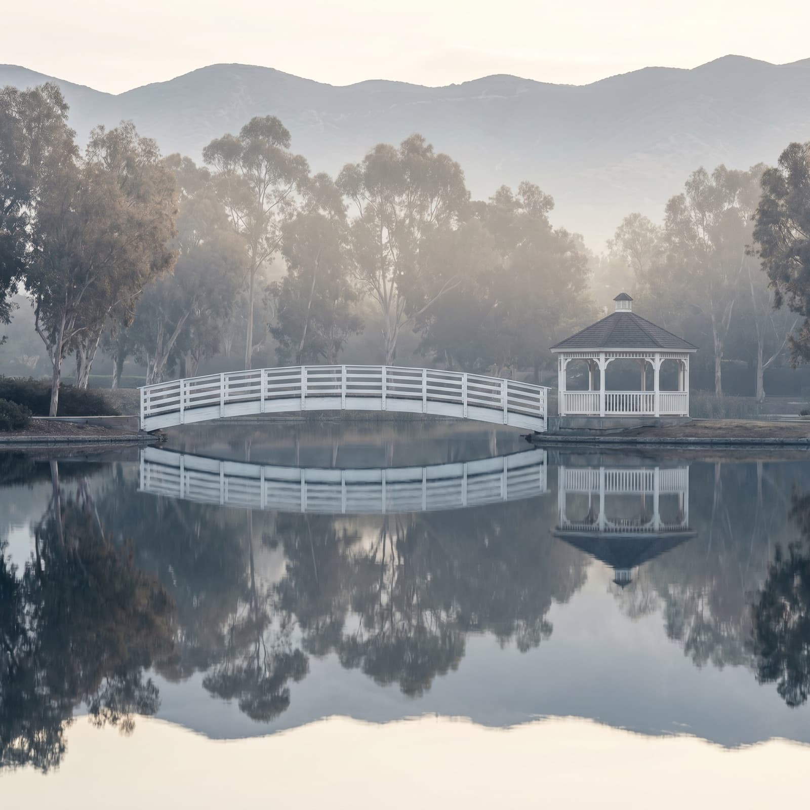 Irvine, CA — The man-made north lake at Woodbridge just after sunrise, the white wooden footbridge and beach-club pavilion reflected dead-still in the water, ducks barely moving, the Santa Ana foothills a soft grey line behind the eucalyptus, the deliberate, planned calm that locals know is the whole point of Irvine.