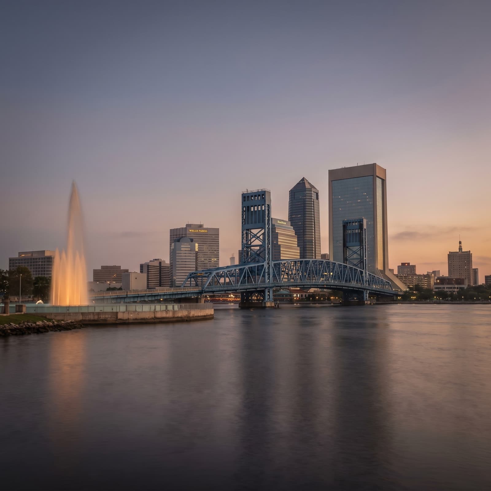 Jacksonville, FL — The Main Street Bridge's blue vertical-lift span over the St. Johns River at dusk, downtown Jacksonville's skyline lit behind it, the Friendship Fountain plume catching the last light on the Southbank, a container ship's silhouette far downriver, no people.