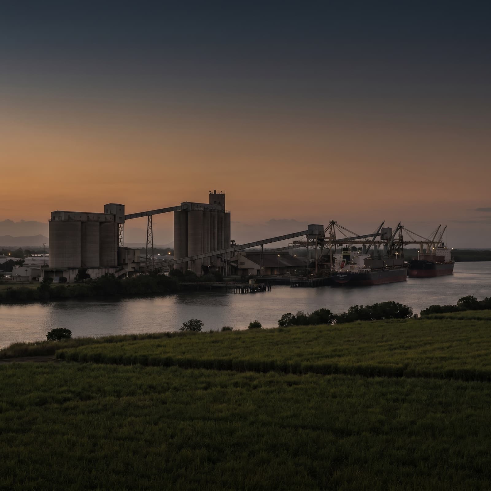 Mackay, QLD — Mackay's working harbour and the bulk-sugar and coal terminals at dusk, cane fields and the Pioneer River in the foreground, industrial and honest, restrained and editorial.