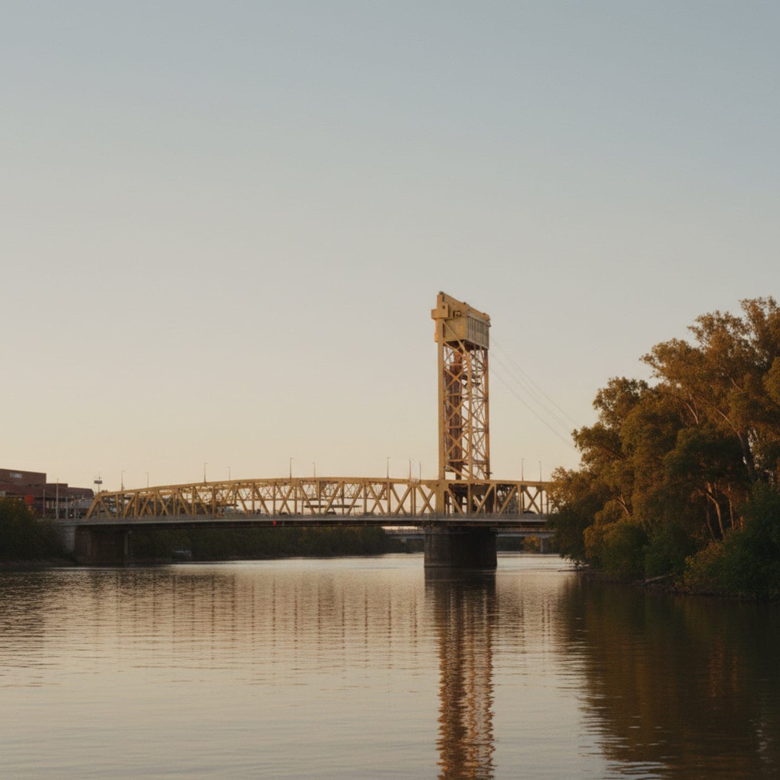 Sacramento, CA — The Tower Bridge over the Sacramento River caught from the West Sacramento side at dusk, the gold paint still warm under the falling light, the Capitol dome small in the distance behind, no joggers on the riverwalk, the river surface glassy and still.