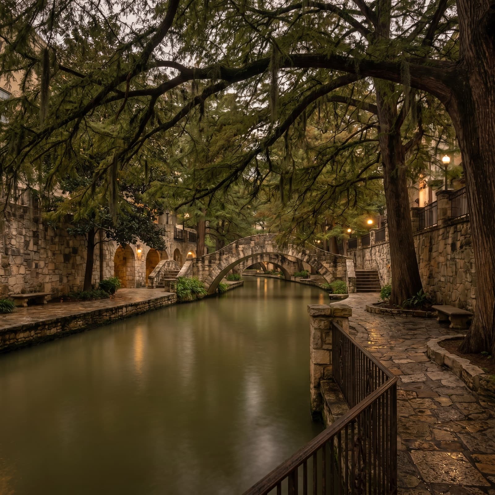 San Antonio, TX — The San Antonio River Walk one level below the street at dusk, cypress branches arching over the green water, the stone footbridges and a lone barge throwing soft light across the cobblestone path, shot from a quiet stretch away from the restaurant crowds, the everyday angle a local takes on the walk back to the car, not the postcard.