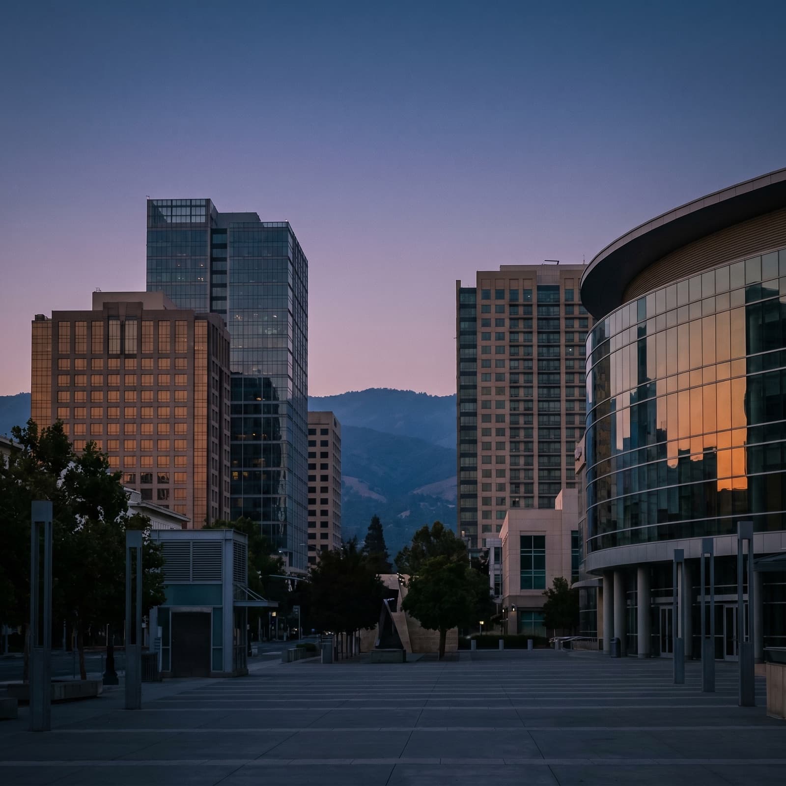 San Jose, CA — Downtown San Jose skyline at dusk from a low vantage near San Pedro Square, the SAP Center's glass curve catching the last light to one side, the foothills of the Diablo Range fading blue behind the towers, warm amber windows against a cooling sky, no people in frame.