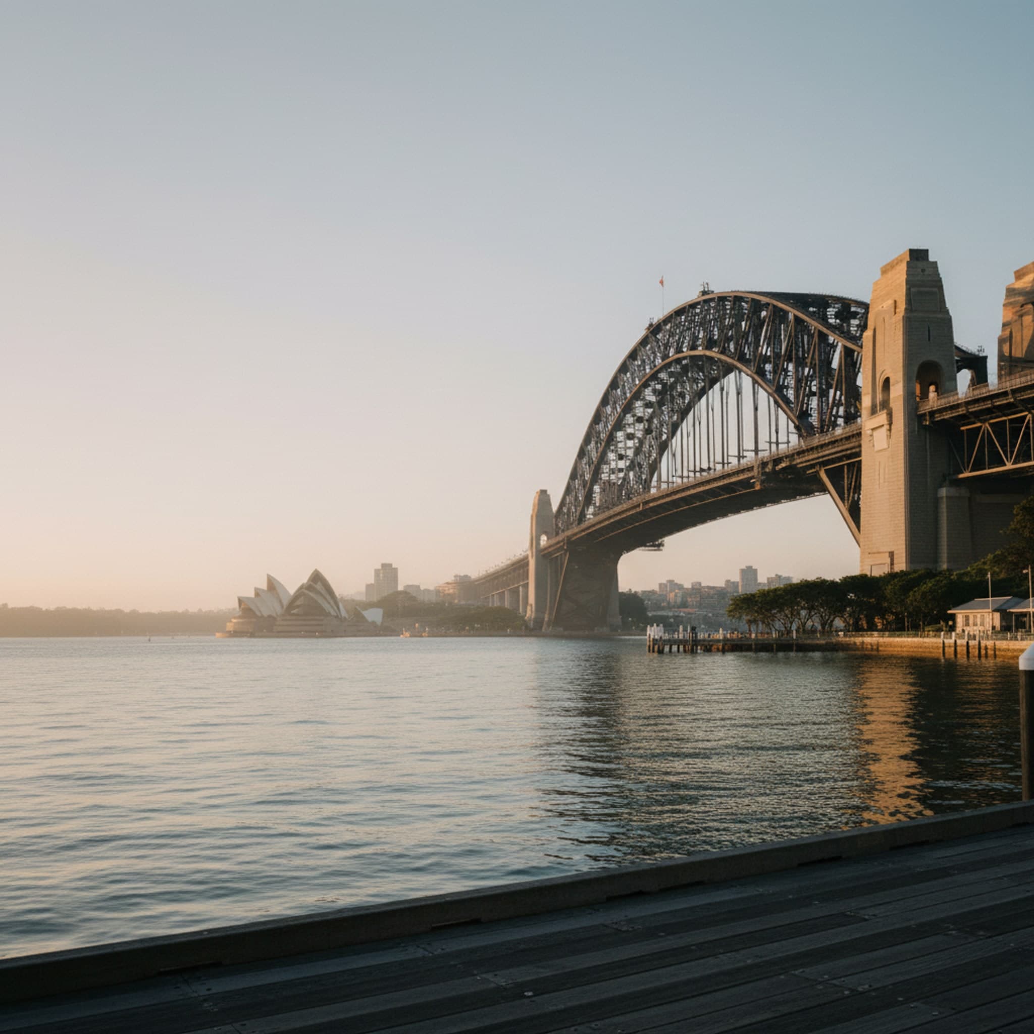 Sydney, NSW — Sydney Harbour seen from a quiet stretch of the eastern foreshore at first light, Harbour Bridge dominating the frame from a low oblique angle, Opera House sails just visible at the right edge in soft cool light. Water glassy and mauve. Restrained, almost editorial — not the postcard angle.