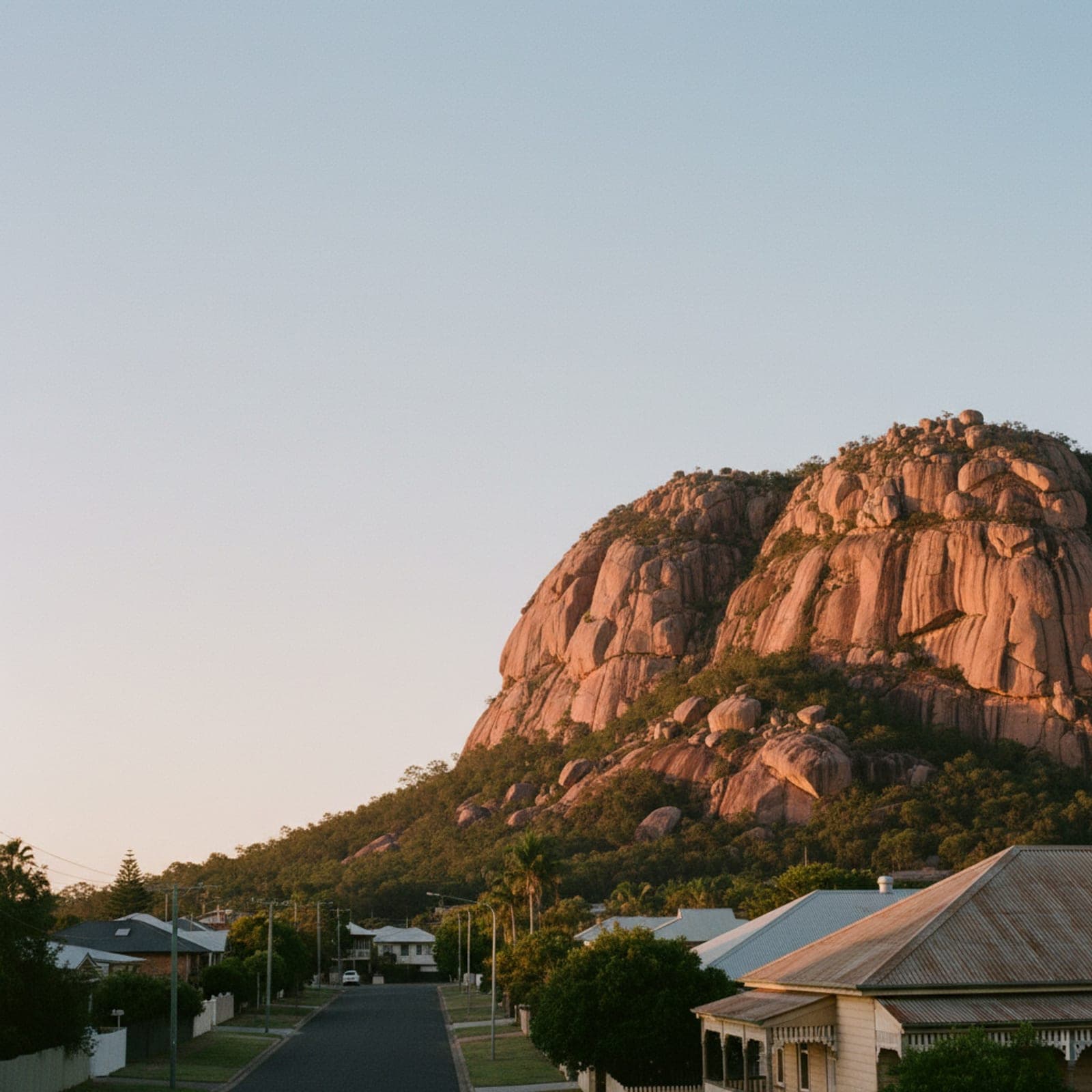 Townsville, QLD — Castle Hill at dawn from a low angle on The Strand, the granite monolith catching warm copper light against a soft tropical sky, Cleveland Bay calm to the right, a single jogger in the foreground for scale. Heat haze just visible, no cliché reef-and-palms shot.